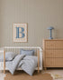 A nursery with a white crib, blue and white striped bedding, Pinstripe Wallpaper Green, a chest of drawers with a blue lamp, a framed letter B on the wall, a round jute rug, and a children&