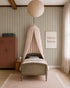 A cozy nursery features a single bed under a blush canopy, a large paper lantern, a pink wardrobe, framed "bisou bisou" art, and Pinstripe Wallpaper Moss adding warmth as sunlight filters through sheer curtains.