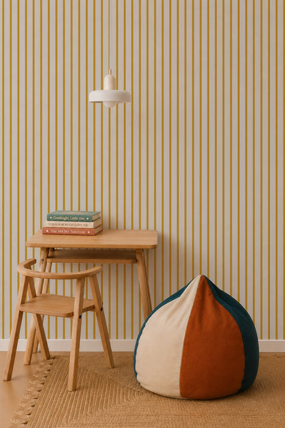 A minimalist room with a small wooden desk and chair, stacked books, a round beanbag in cream, orange, and green, and a hanging white lamp set against Pinstripe Wallpaper Mustard.