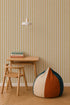 A minimalist room with a small wooden desk and chair, stacked books, a round beanbag in cream, orange, and green, and a hanging white lamp set against Pinstripe Wallpaper Mustard.