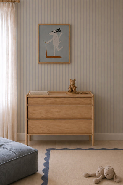 A light wood dresser with a teddy bear and book stands against Pinstripe Wallpaper Sky in the nursery. Above it hangs a framed dog drawing, while plush toys and a blue cushion rest on a scalloped rug by flowing curtains.
