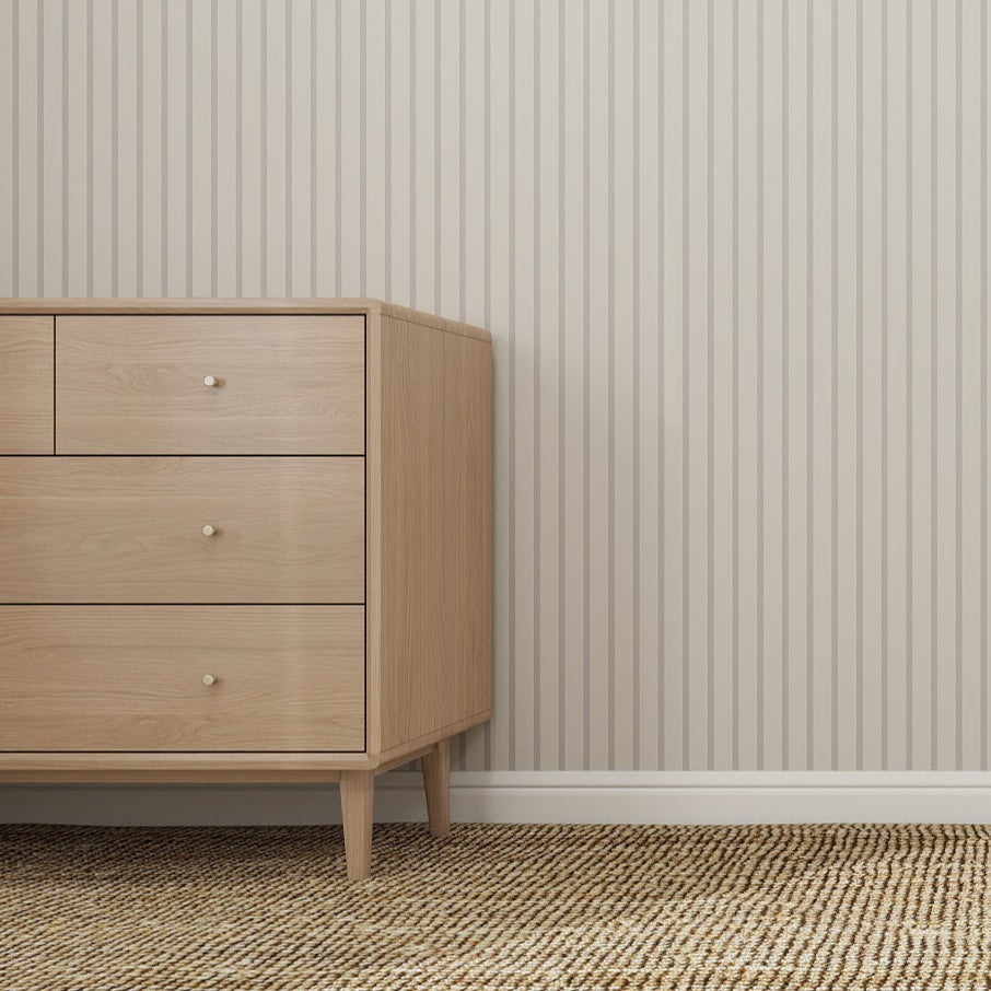 A light wooden dresser stands on the left side of a room featuring Pinstripe Wallpaper Stone, a woven rug, and sunlight streaming in from a window out of frame.