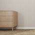 A light wooden dresser stands on the left side of a room featuring Pinstripe Wallpaper Stone, a woven rug, and sunlight streaming in from a window out of frame.