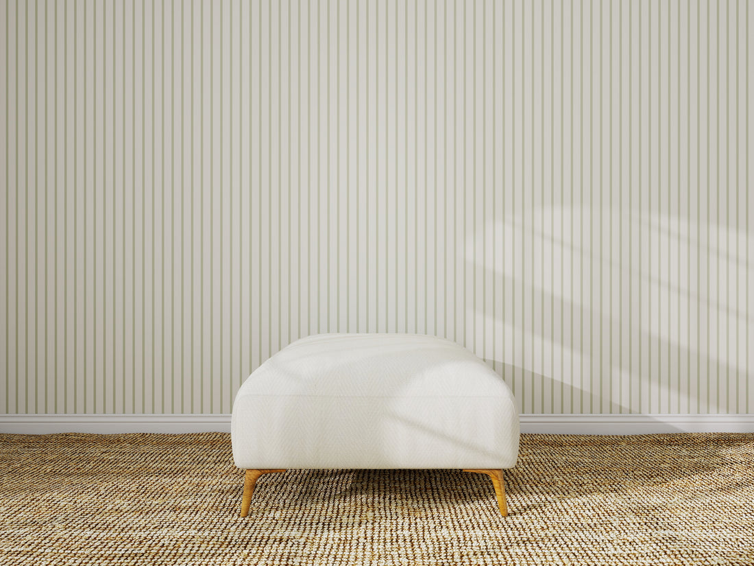 A minimalist room features a Pinstripe Green wallpaper sample on the wall, a textured tan carpet, and a single white ottoman with wooden legs in the center. Sunlight casts gentle shadows on the wall.