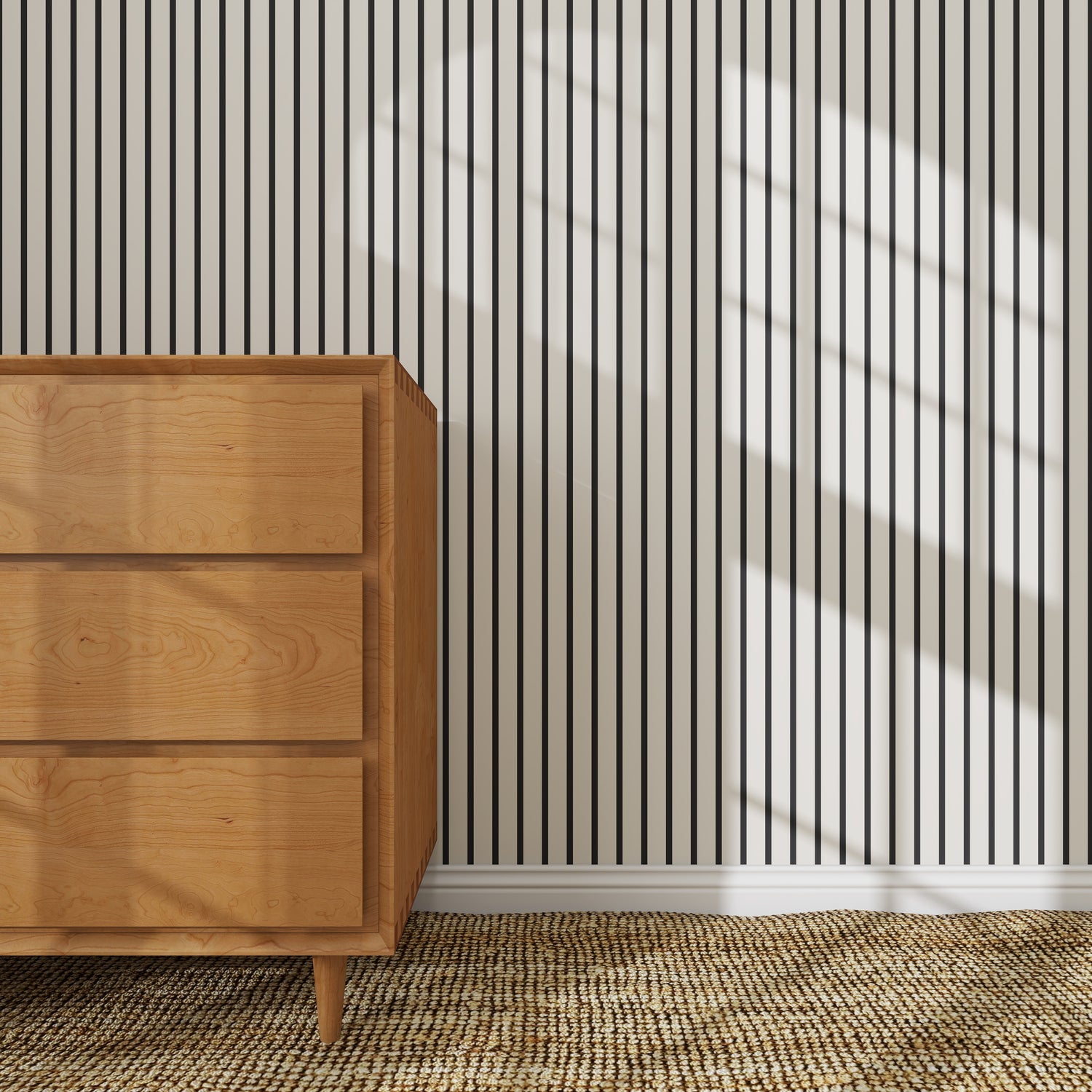 A wooden dresser sits on a woven rug before the Pinstripe Wallpaper Sample, Charcoal. Sunlight streams in, casting the arched shadow of a large window across both the wall and floor.