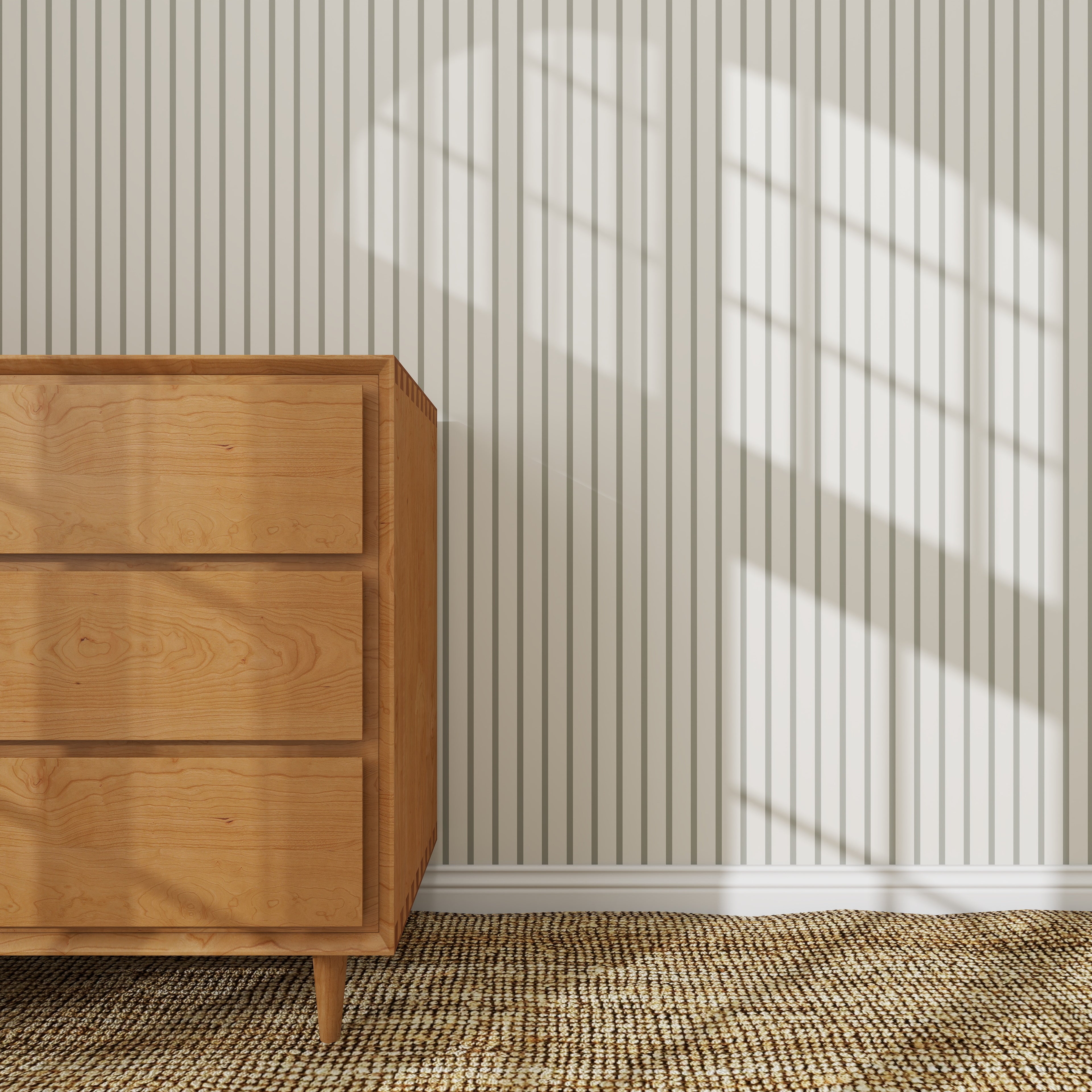 A wooden dresser stands on a woven rug by a white wall with Pinstripe Wallpaper Matcha. Sunlight streams through the window, casting window pane shadows on the striped wallpapered wall.