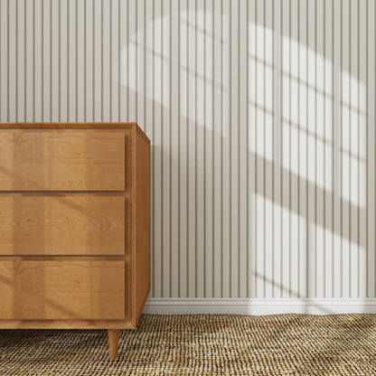 A wooden dresser stands on a woven rug by a white wall with Pinstripe Wallpaper Matcha. Sunlight streams through the window, casting window pane shadows on the striped wallpapered wall.