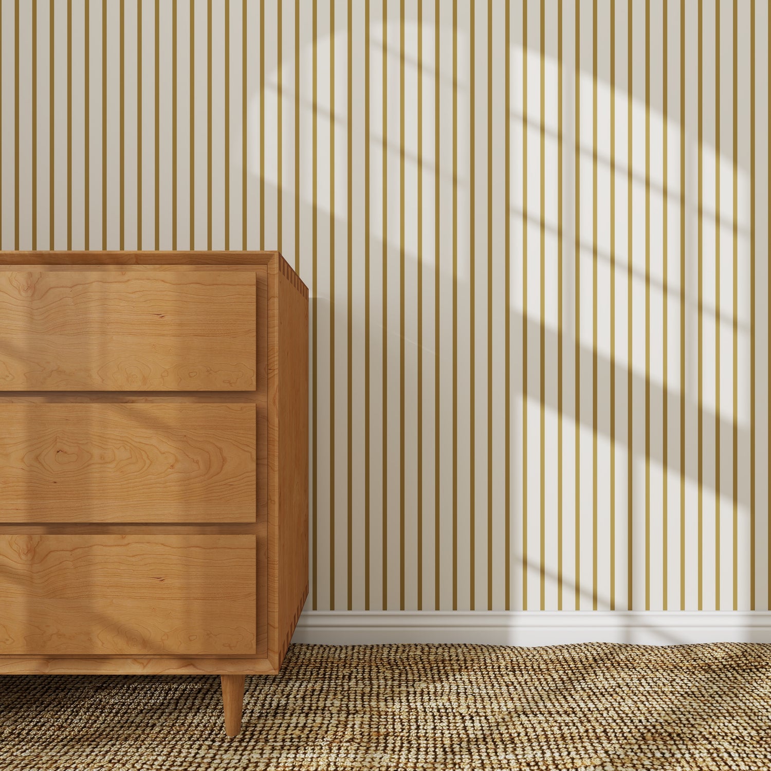 A wooden dresser stands on a woven rug against a wall decorated with Pinstripe Wallpaper Sample, Moss, featuring beige and brown vertical stripes. Sunlight casts window-shaped shadows, creating a bright, cozy atmosphere.