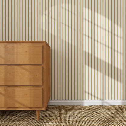 A wooden dresser stands on a woven rug against a wall decorated with Pinstripe Wallpaper Sample, Moss, featuring beige and brown vertical stripes. Sunlight casts window-shaped shadows, creating a bright, cozy atmosphere.