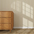A wooden dresser stands on a woven rug against a wall decorated with Pinstripe Wallpaper Sample, Moss, featuring beige and brown vertical stripes. Sunlight casts window-shaped shadows, creating a bright, cozy atmosphere.