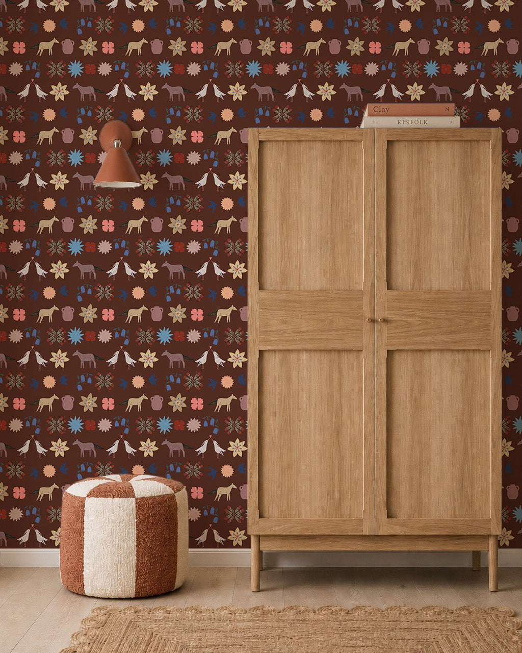A wooden wardrobe stands on a woven rug beside a round, red and white pouf. The dark brown wall is decorated with Playful Prairie Cocoa Wallpaper, featuring whimsical animal and floral patterns. A small wall lamp is mounted above the pouf.