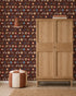A wooden wardrobe stands on a woven rug beside a round, red and white pouf. The dark brown wall is decorated with Playful Prairie Cocoa Wallpaper, featuring whimsical animal and floral patterns. A small wall lamp is mounted above the pouf.