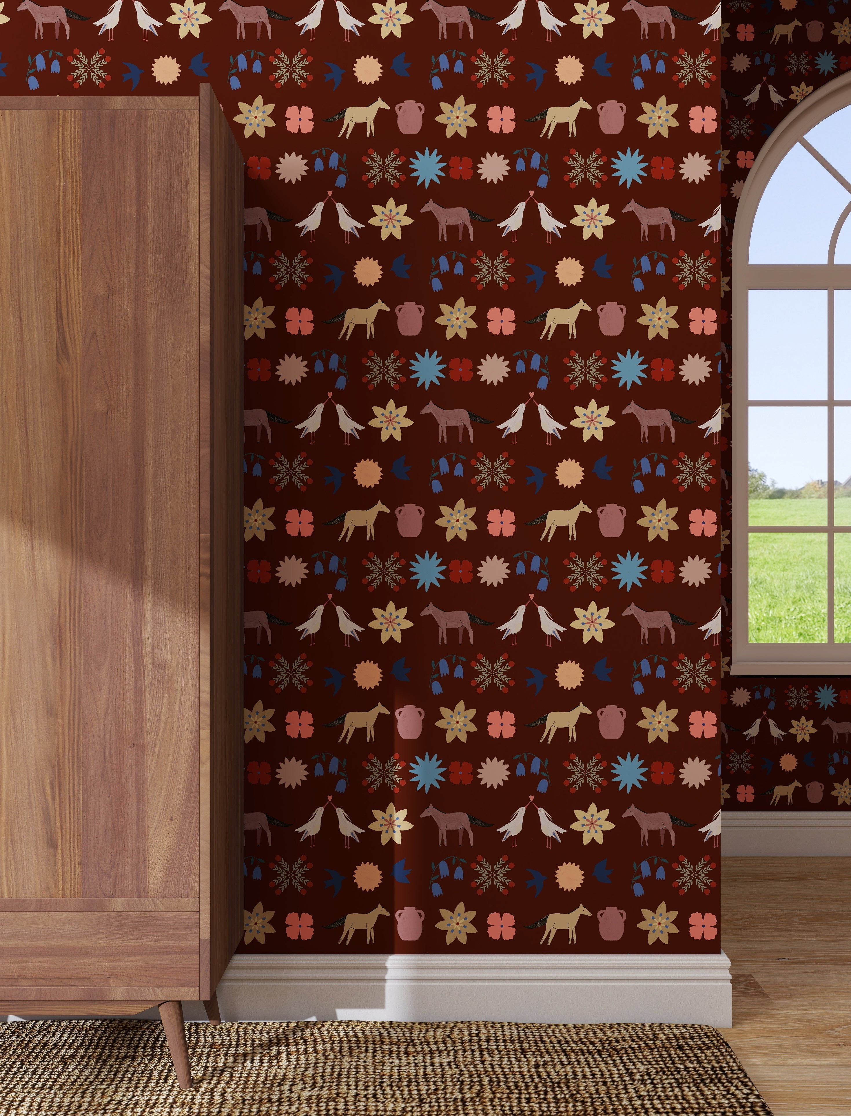 A wooden wardrobe stands on a woven rug by a maroon wall decorated with the Playful Prairie Cocoa Wallpaper Sample, which features folk-style horses, birds, flowers, and plants. A window offers a view of grassy outdoors.