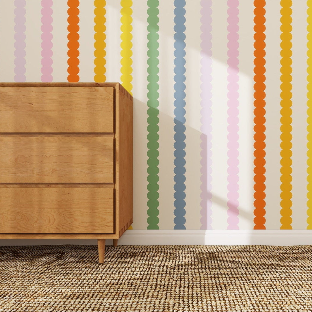 A wooden dresser sits on a rug beside a wall decorated with Scallop Stripe Wallpaper Repeat Pattern | Rainbow, featuring circular shapes in orange, green, blue, yellow, pink, and red. Sunlight highlights the playful design.