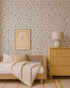 A cozy children’s bedroom features Powdered Petals Wallpaper, a wooden bed with striped bedding, a yellow dresser, matching lamp, and a framed bunny illustration in soft neutral and pastel tones.