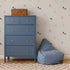 A blue dresser with a wicker basket on top sits next to a blue bean bag chair. The wall is decorated with Puppy Dog Wallpaper, perfect for dog lovers’ decor, while a beige checkered rug covers the wooden floor.