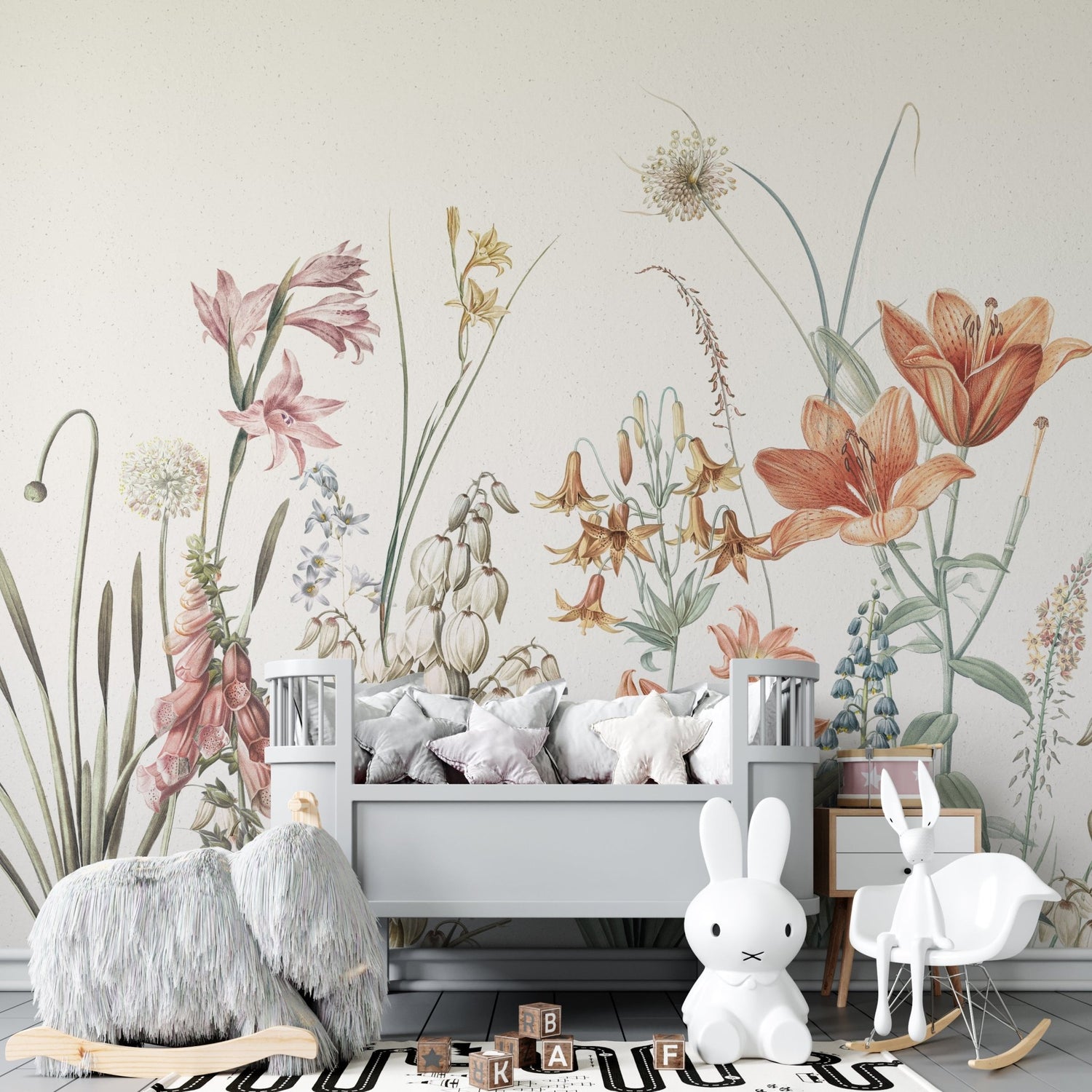 A nursery with a gray crib, plush cushions, and a black-and-white rug. The Quinns Flower Field Mural Sample adds floral flair to the wall, while a white bunny lamp and furry animal seat sit by the crib; wooden blocks rest on the rug.
