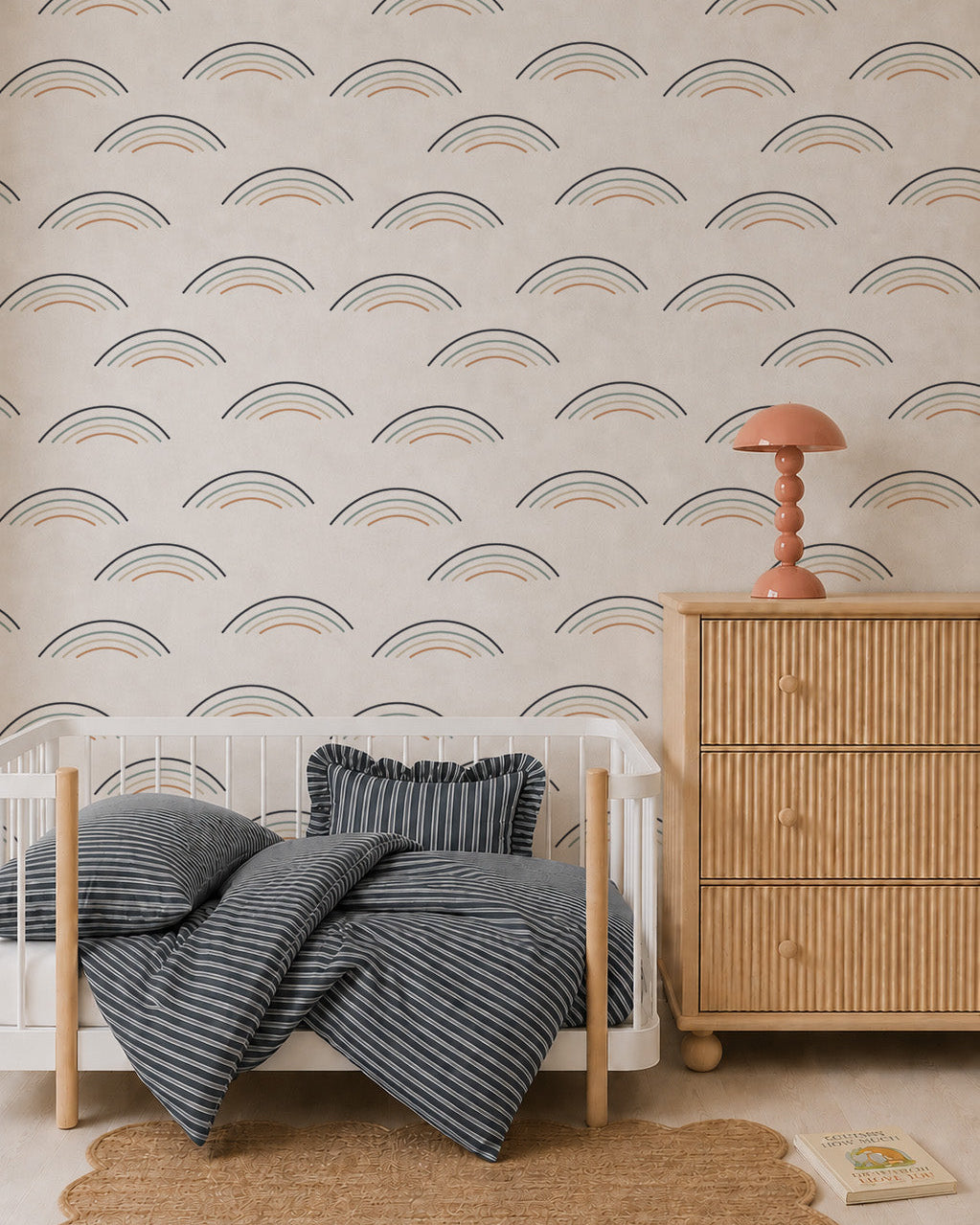 A cozy nursery with a white crib, blue striped bedding and pillow, rattan dresser with a coral lamp, scalloped rug, Astrid Unicorn Wallpaper, and a children’s book on the floor.