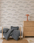 A cozy nursery with a white crib, blue striped bedding and pillow, rattan dresser with a coral lamp, scalloped rug, Astrid Unicorn Wallpaper, and a children’s book on the floor.