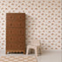 A tall brown dresser stands next to a small beige child’s chair on a light rug. The background wall features repeating pattern wallpaper adorned with simple orange rainbows, identified as the Raining Rainbows Wallpaper.