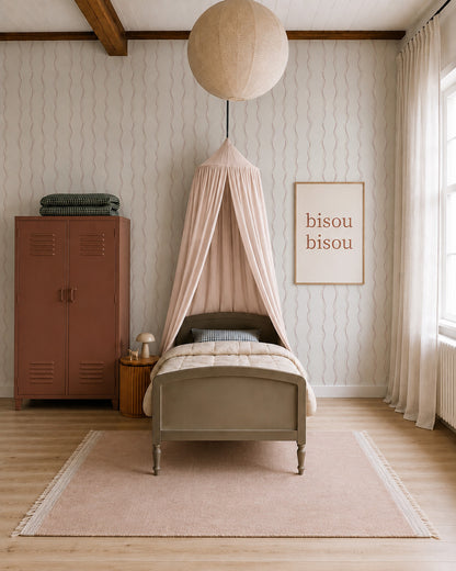 A cozy bedroom with a single bed under a blush canopy, a round ceiling light, a pink locker, a small side table, and a framed bisou bisou print on Ribbon Stripe Wallpaper in Rose. Light streams in from a large window, highlighting the charming nursery wallpaper touches throughout the space.