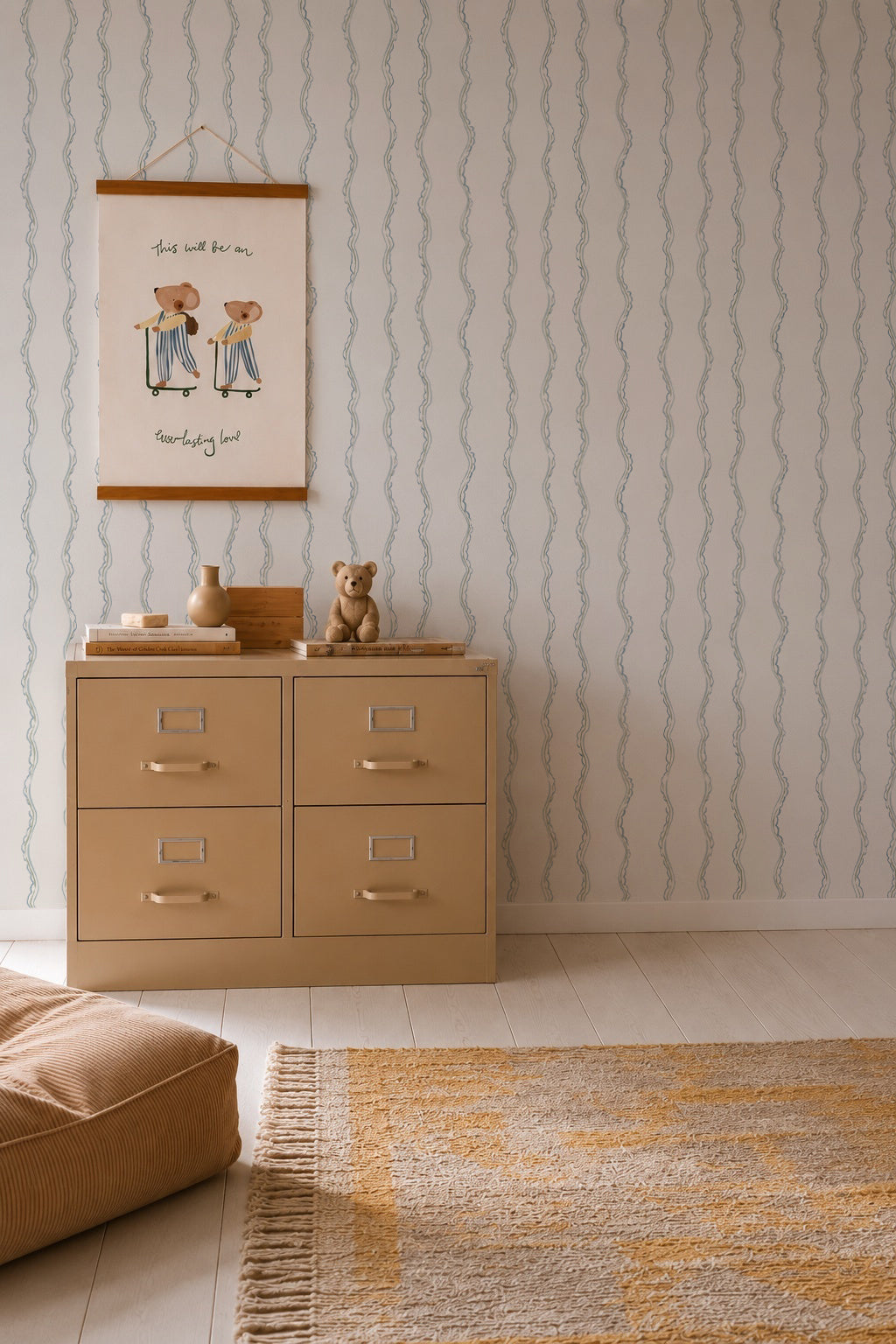 A beige dresser with four drawers stands against Ribbon Stripe Wallpaper in blue. On top are a framed drawing, toys, and a teddy bear. A cushioned seat and a yellow scallop-striped woven rug complete the calming nursery decor on the light wood floor.