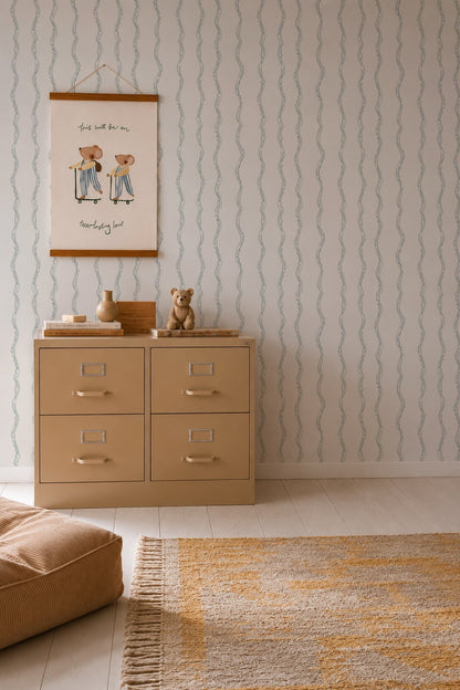 A beige dresser with four drawers stands against Ribbon Stripe Wallpaper in blue. On top are a framed drawing, toys, and a teddy bear. A cushioned seat and a yellow scallop-striped woven rug complete the calming nursery decor on the light wood floor.