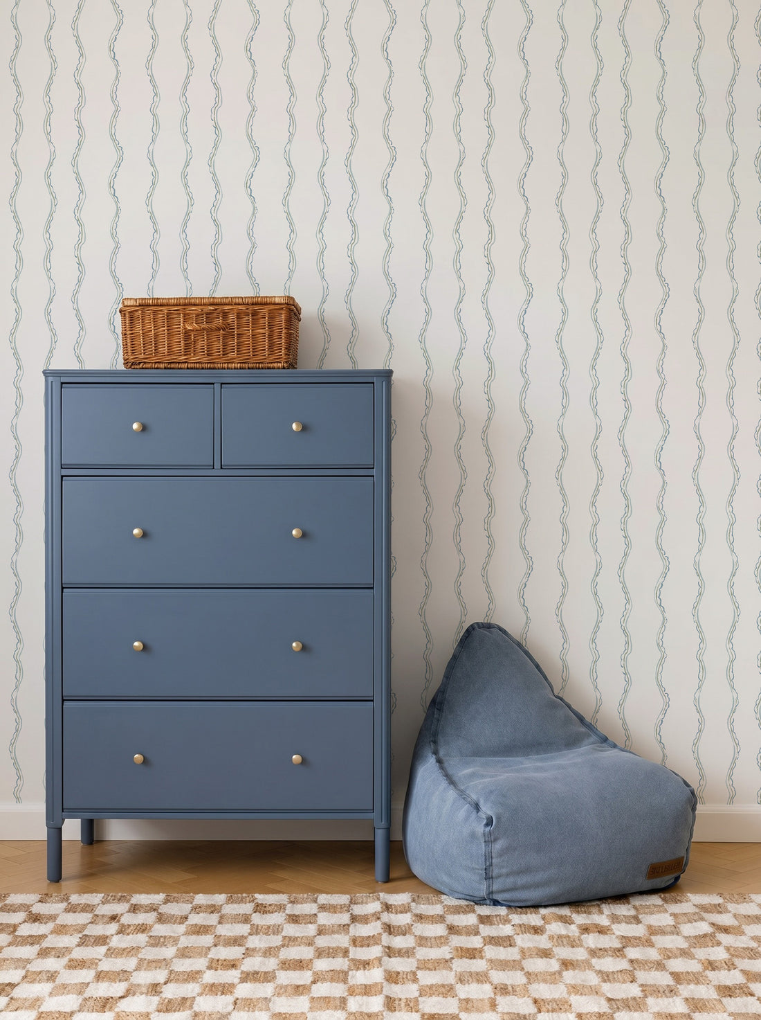 A blue dresser with four drawers and a wicker basket on top stands next to a blue bean bag chair. The background features Ribbon Stripe Wallpaper in Blue and a beige checkered rug, creating calming nursery decor.