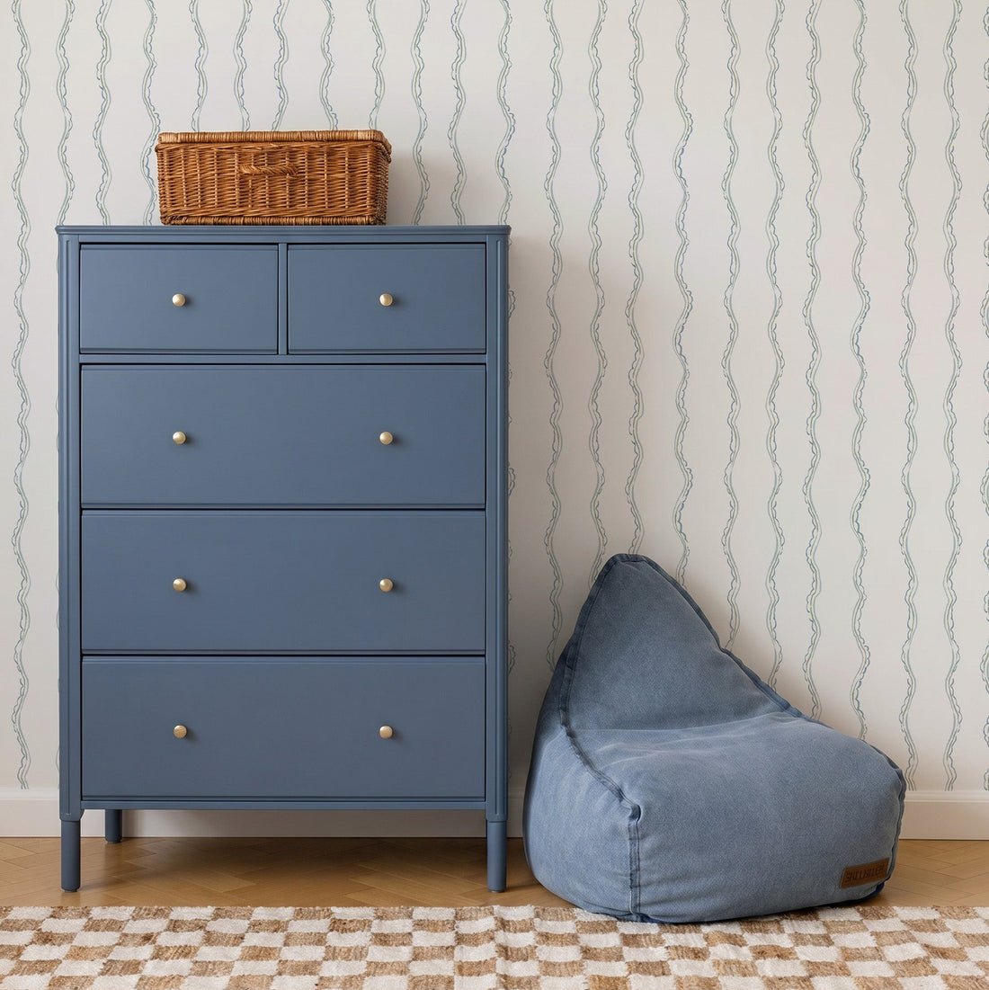 A blue dresser with four drawers and a wicker basket on top stands next to a blue bean bag chair. The background features Ribbon Stripe Wallpaper in Blue and a beige checkered rug, creating calming nursery decor.