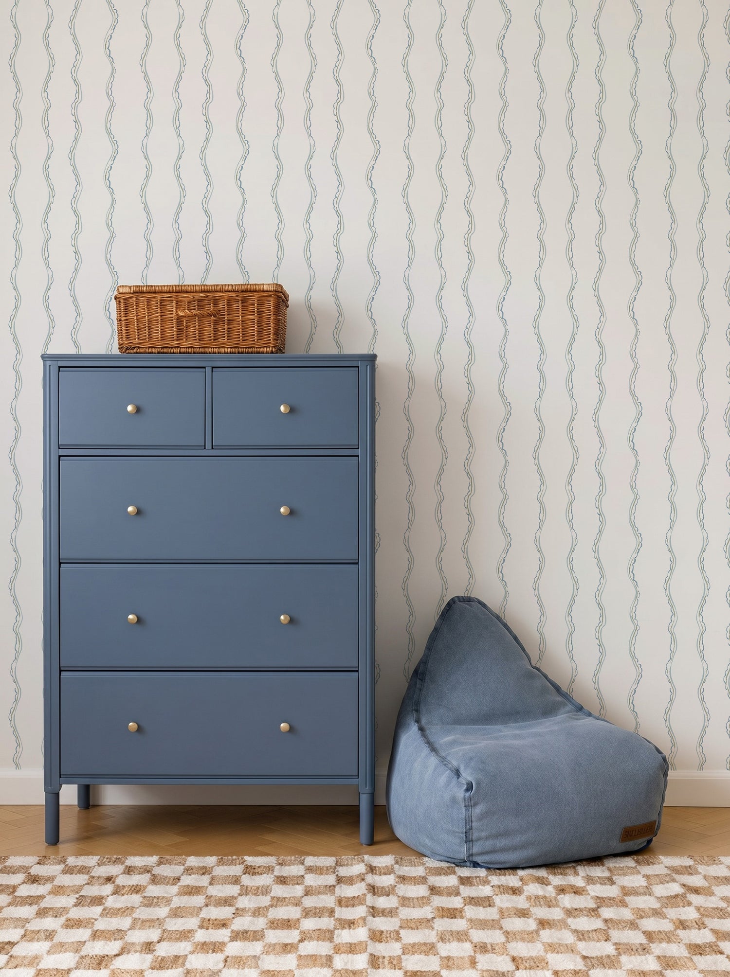 A blue dresser with four drawers and a wicker basket on top stands next to a blue bean bag chair. The background features Ribbon Stripe Wallpaper in Blue and a beige checkered rug, creating calming nursery decor.