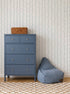 A blue dresser with four drawers and a wicker basket on top stands next to a blue bean bag chair. The background features Ribbon Stripe Wallpaper in Blue and a beige checkered rug, creating calming nursery decor.