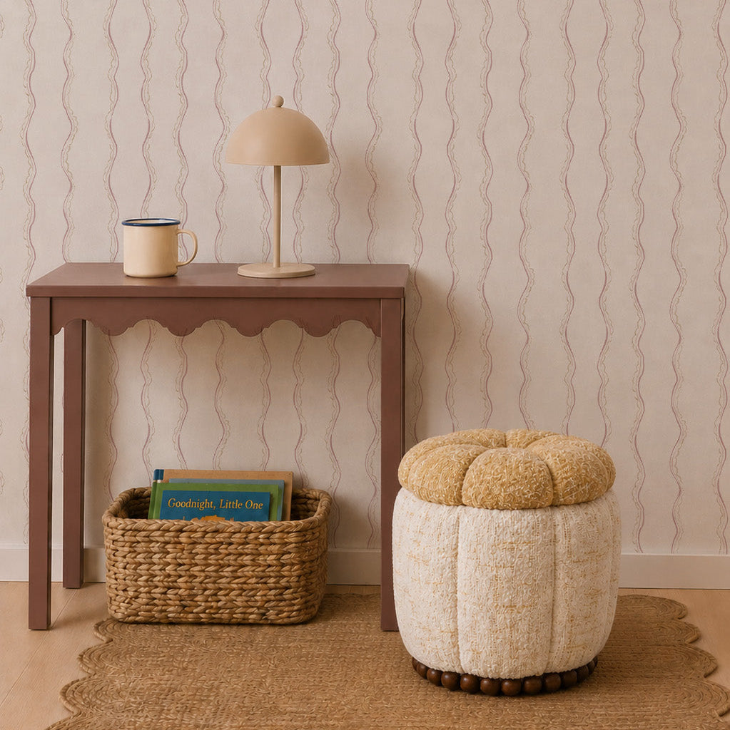 A small brown table with a lamp and mug, a woven basket filled with books, and a round beige ottoman rest on a woven rug against Ribbon Stripe Wallpaper in Rose, creating an inviting atmosphere accented by the wavy-patterned background.