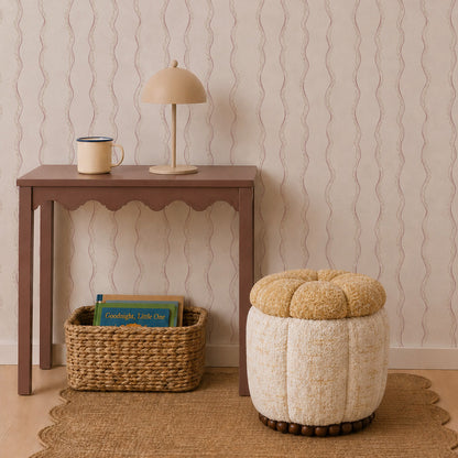 A small brown table with a lamp and mug, a woven basket filled with books, and a round beige ottoman rest on a woven rug against Ribbon Stripe Wallpaper in Rose, creating an inviting atmosphere accented by the wavy-patterned background.