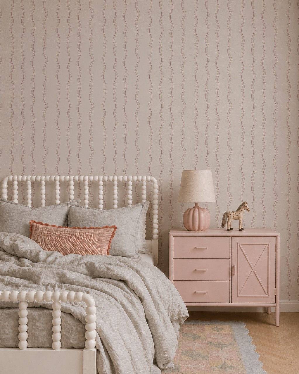 A cozy bedroom with a white spindle bed, gray and blush bedding, a light pink dresser, pink lamp, toy horse, pastel rug, and Ribbon Stripe Wallpaper in Rose for a charming touch with its subtle scallop stripe design.