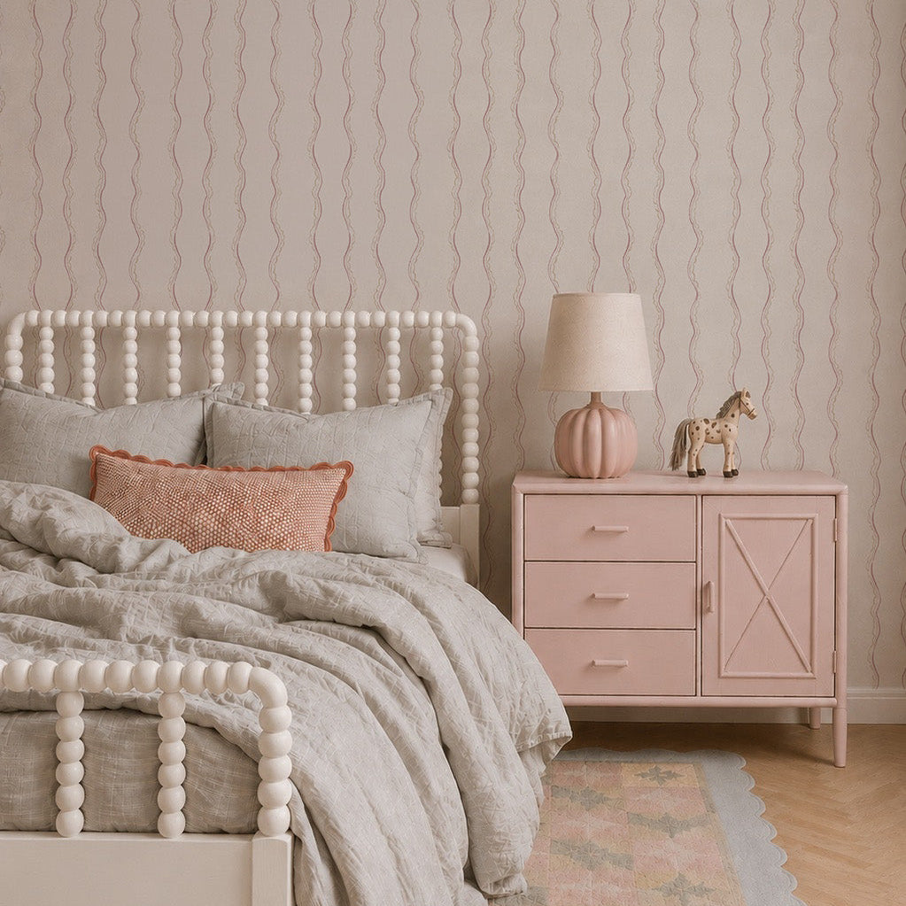 A cozy bedroom features a white spindle bed with light gray bedding, a coral pillow, and a pink nightstand topped with a lamp and giraffe figurine. A pastel rug lies on the floor beside a children’s book, while the space is elegantly accented by Ribbon Stripe Wallpaper in Rose adorning the walls.