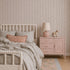 A cozy bedroom features a white spindle bed with light gray bedding, a coral pillow, and a pink nightstand topped with a lamp and giraffe figurine. A pastel rug lies on the floor beside a children’s book, while the space is elegantly accented by Ribbon Stripe Wallpaper in Rose adorning the walls.