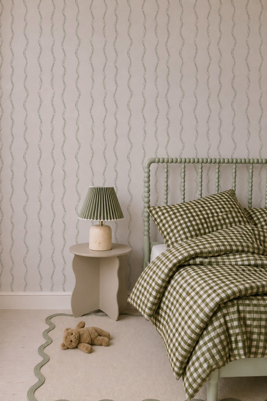 A cozy bedroom features a green spindle bed, green and white checkered bedding, a small beige nightstand with a green lamp, a wavy-edged beige rug, a teddy bear on the floor, and playful charm from Ribbon Stripe Wallpaper in Sage.