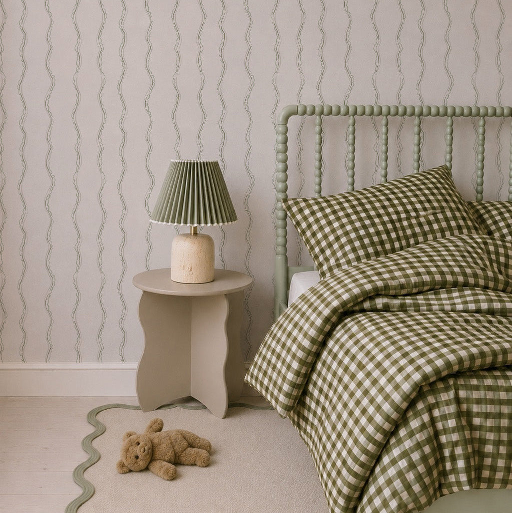 A cozy bedroom features a green spindle bed, green and white checkered bedding, a small beige nightstand with a green lamp, a wavy-edged beige rug, a teddy bear on the floor, and playful charm from Ribbon Stripe Wallpaper in Sage.