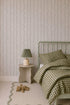 A cozy bedroom features a green spindle bed, green and white checkered bedding, a small beige nightstand with a green lamp, a wavy-edged beige rug, a teddy bear on the floor, and playful charm from Ribbon Stripe Wallpaper in Sage.