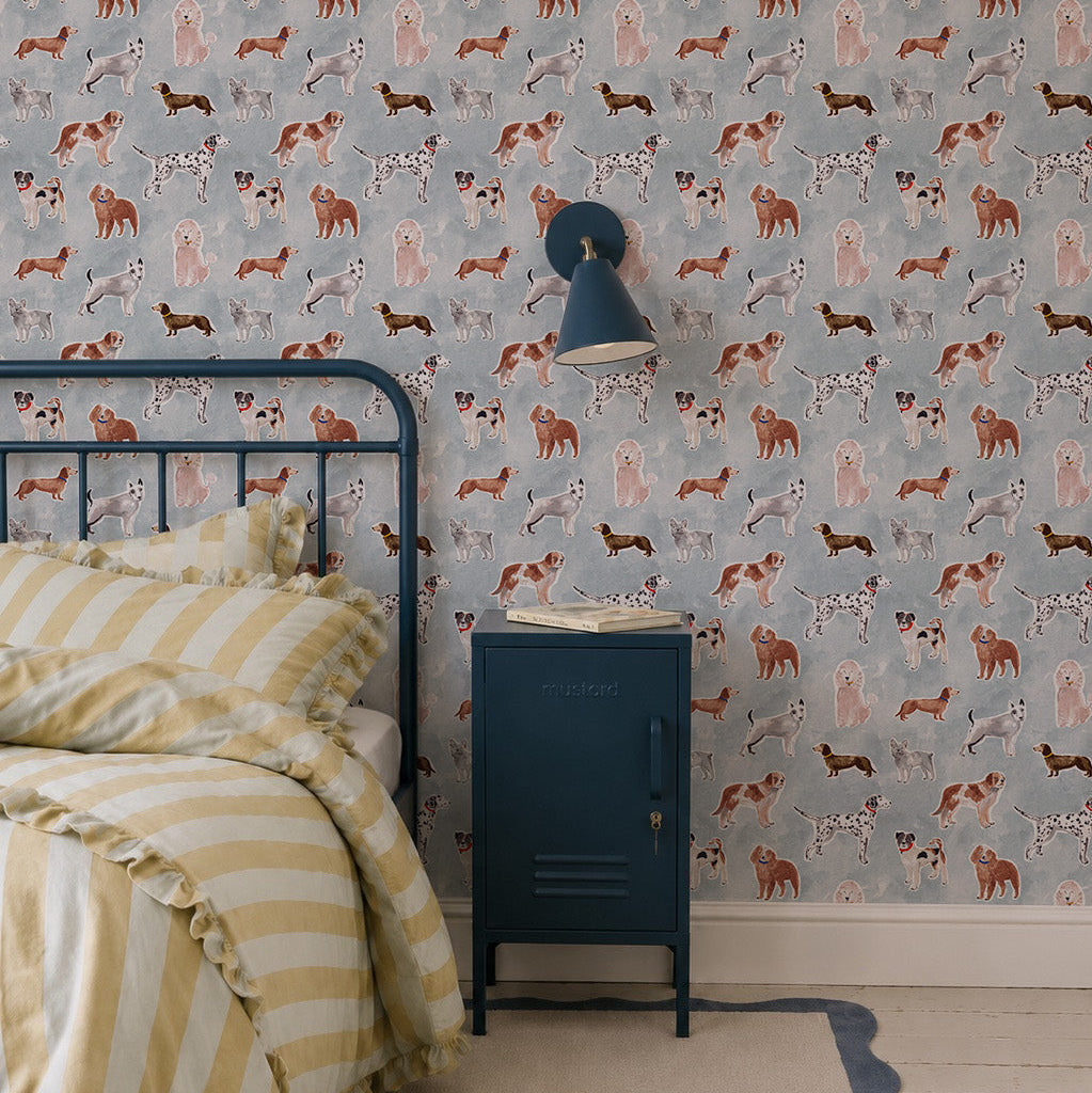 A cozy bedroom features a metal-framed bed, yellow and white striped bedding, a blue nightstand, a wall lamp, and cheerful Rigby Dog Print Wallpaper in blue on a gray background.