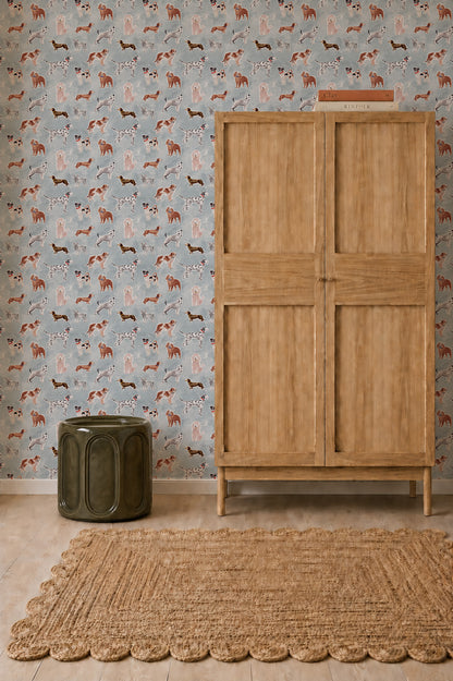 Rigby Dog Print Wallpaper, Blue featuring a repeating pattern of various dog breeds, shown behind a wooden armoire, a dark green stool, and a woven rug.