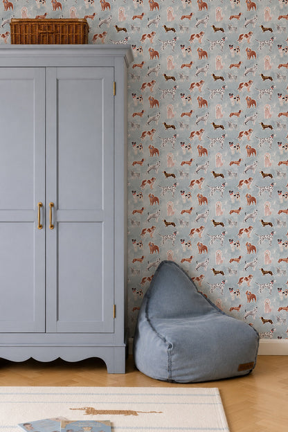 Rigby Dog Print Wallpaper, Blue featuring a repeating pattern of various dog breeds, shown behind a grey wardrobe and a floor cushion.