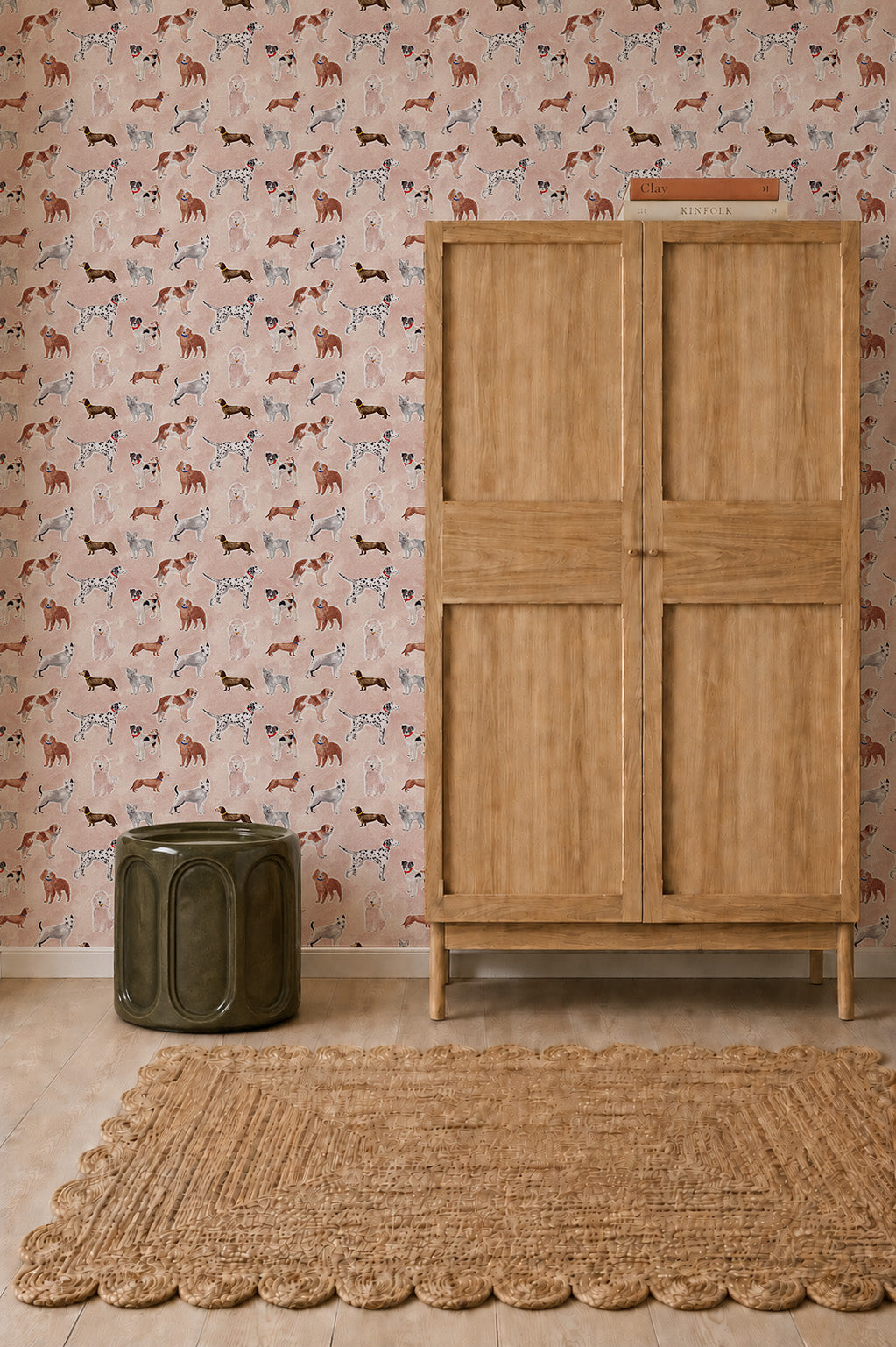 Rigby Dog Print Wallpaper, Rose, features handpainted dogs on a rose-colored background behind a wooden cabinet, a green ceramic stool, and a woven rug.