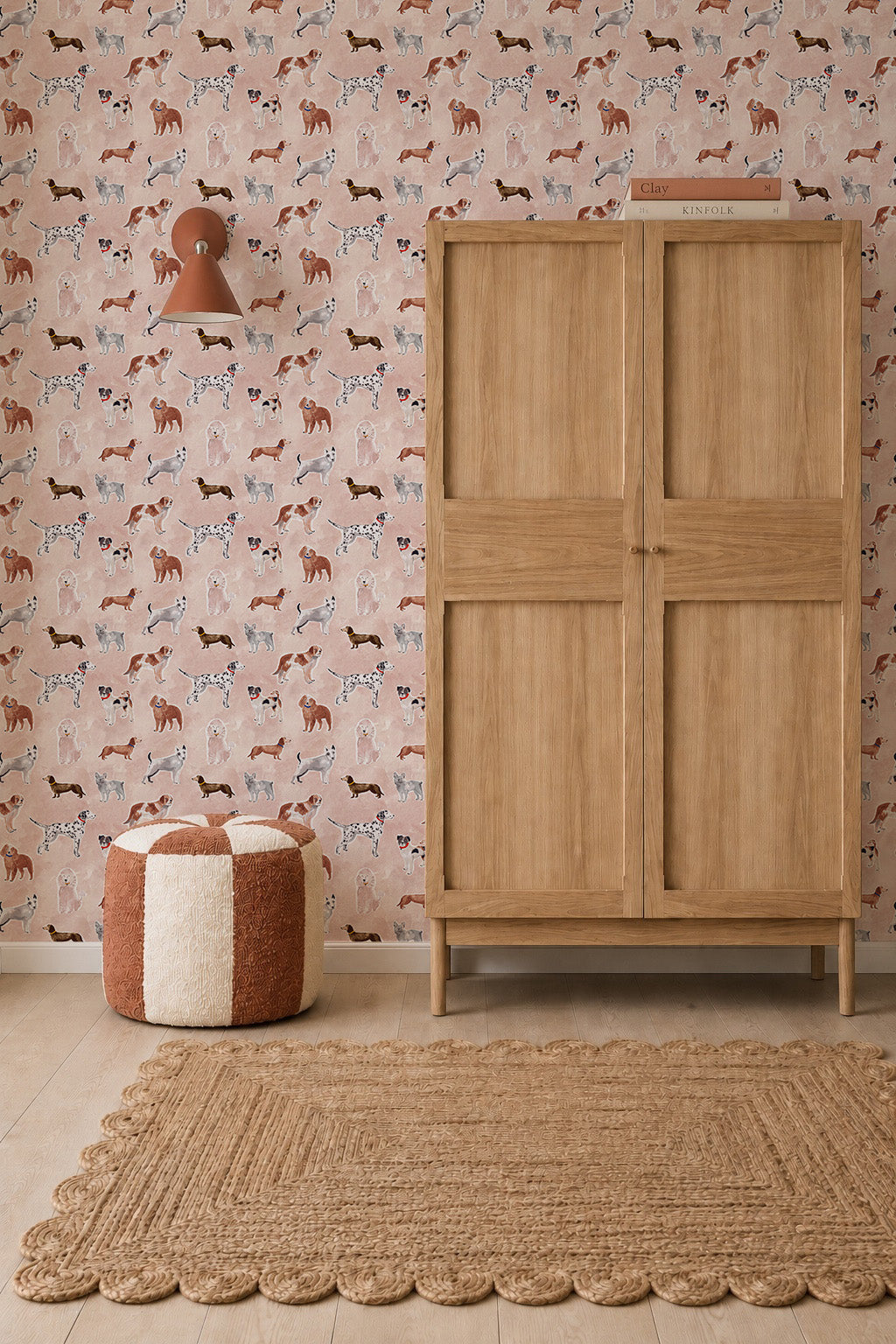 Rigby Dog Print Wallpaper, Rose, featuring a handpainted repeating pattern of various dog breeds on a rose-colored background, shown in a room with a wooden wardrobe, a patterned pouf, and a jute rug.