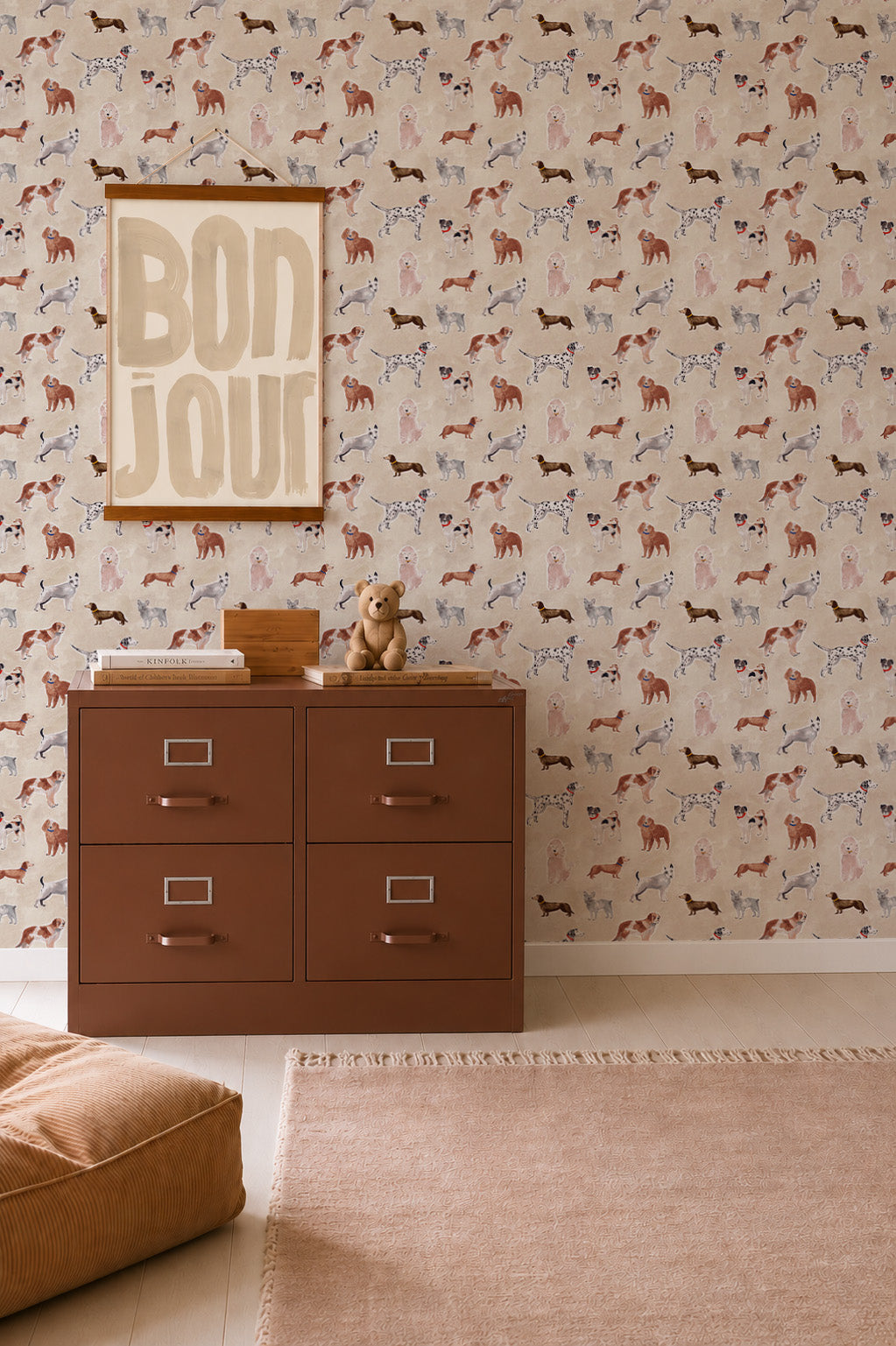 Rigby Dog Print Wallpaper, Sand, featuring a repeating pattern of handpainted dog prints on a sand-colored background, behind a brown metal filing cabinet topped with a teddy bear and books.