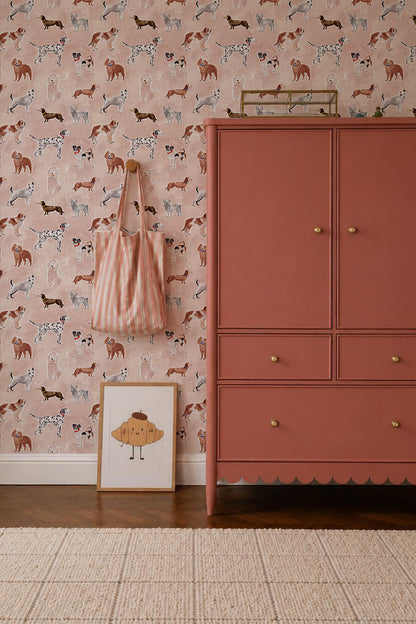 A pink wardrobe stands by a wall covered in Rigby Dog Print Wallpaper, Rose. A striped bag hangs nearby, and a framed muffin illustration rests on the light rug.