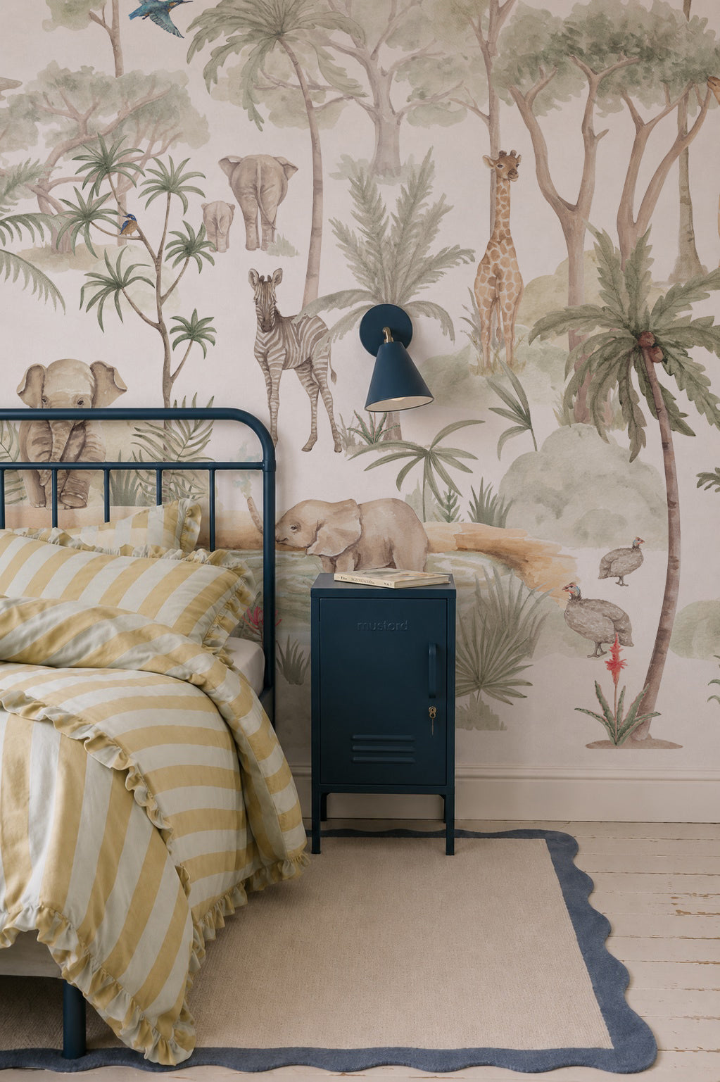 A cozy safari nursery with a metal bed, striped yellow bedding, navy side table with wall lamp, and the Safari Savannah Adventure Mural featuring elephants, giraffes, zebras, palm trees, and birds.