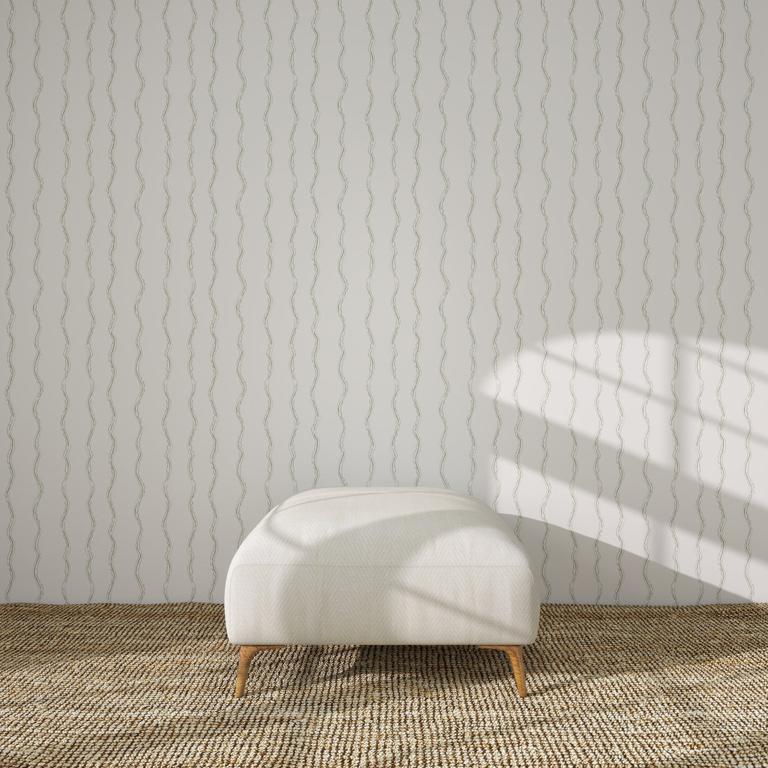 A modern white ottoman with wooden legs sits on a woven beige rug in front of a light gray wall decorated with Ribbon Stripe Wallpaper in Sage. Sunlight and shadows fall across the wall from the right.