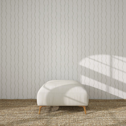 A modern white ottoman with wooden legs sits on a woven beige rug in front of a light gray wall decorated with Ribbon Stripe Wallpaper in Sage. Sunlight and shadows fall across the wall from the right.