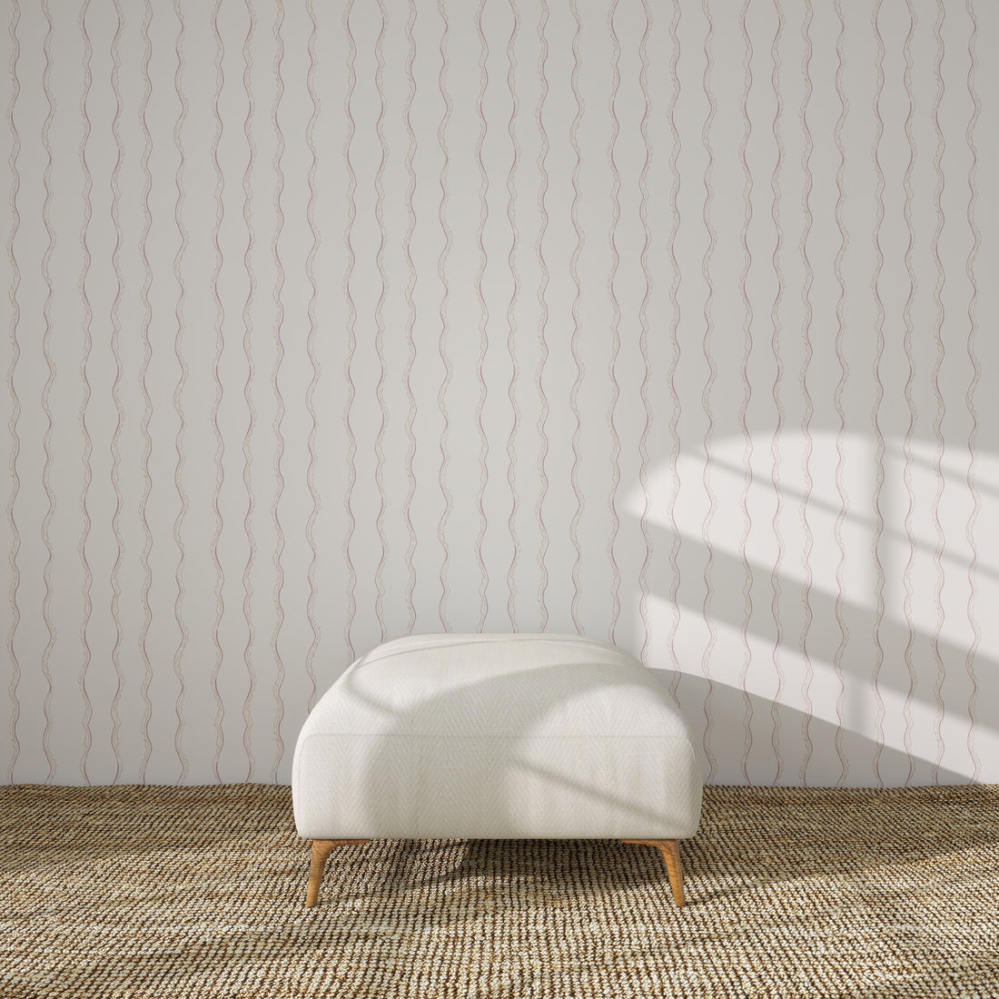 A white upholstered bench with wooden legs sits on a woven rug, against a white wall adorned with Ribbon Stripe Wallpaper in Rose. Sunlight casts gentle shadows, creating a serene scene perfect for elegant children&